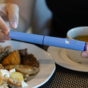 Close-up of hands holding an Ozempic injection pen while sitting at a table with a plate of food and a cup of soup