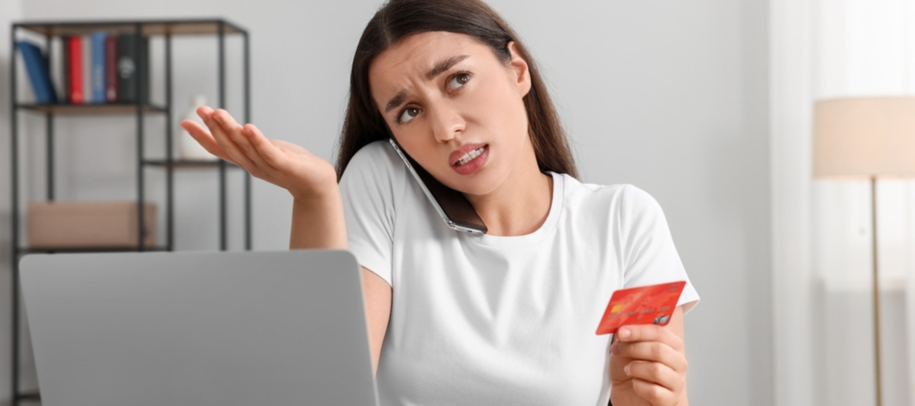Confused woman with credit card talking on phone in front of laptop at table indoors.