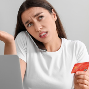 Confused woman with credit card talking on phone in front of laptop at table indoors.