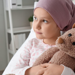 Vials and syringe with Moderna logo + Child undergoing cancer treatment hugs a teddy-bear