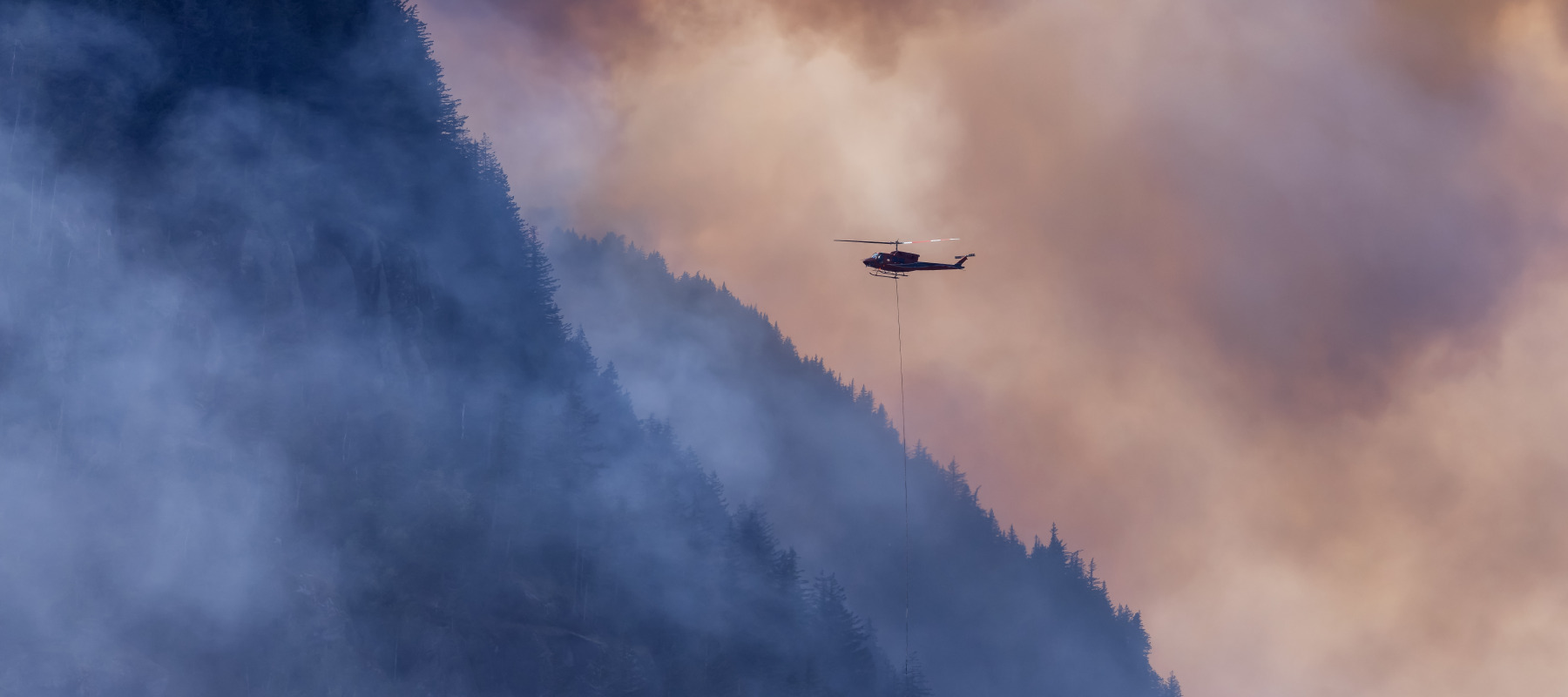 Wildfire Service Helicopter flying over BC Forest Fire and Smoke on the mountain near Hope during a hot sunny summer day