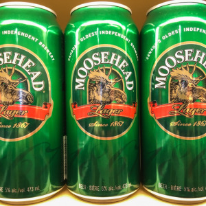 Cans of Moosehead Lager on a retail liquor store shelf + US and Canadian flag near the border symbolizing trade war