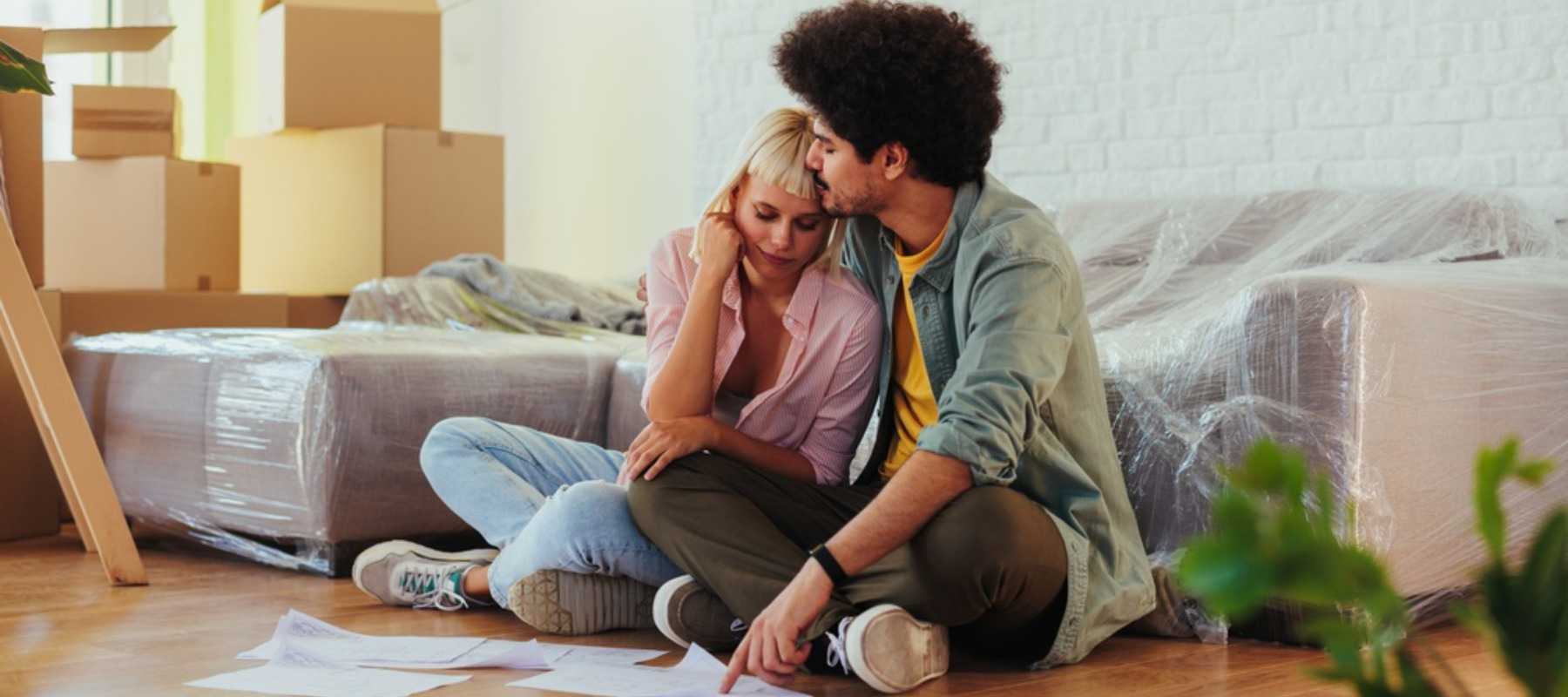 Young couple sitting on floor, surrounded by moving boxes and covered furniture, experiencing stress while planning their new home