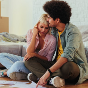 Young couple sitting on floor, surrounded by moving boxes and covered furniture, experiencing stress while planning their new home