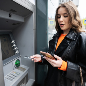 Log in screen concept + Young woman at an ATM looking shocked at her phone